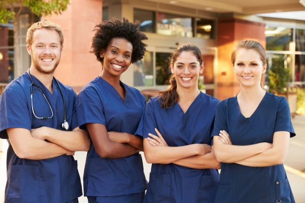 A group of ethnically diverse nurses in blue scrubs smiling outdoors with crossed arms
