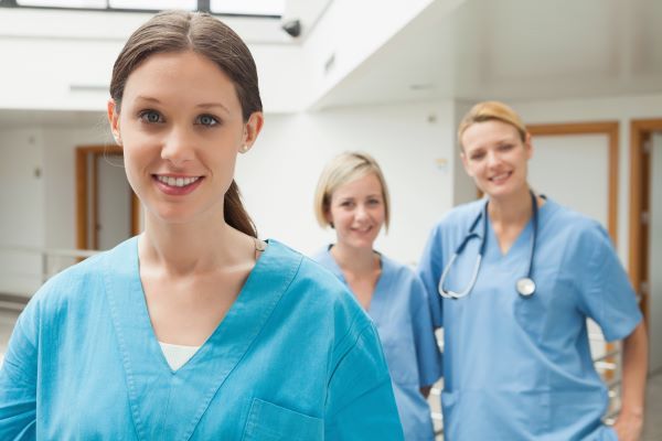 Three smiling nurses wearing scrubs and standing in a hospital corridor
