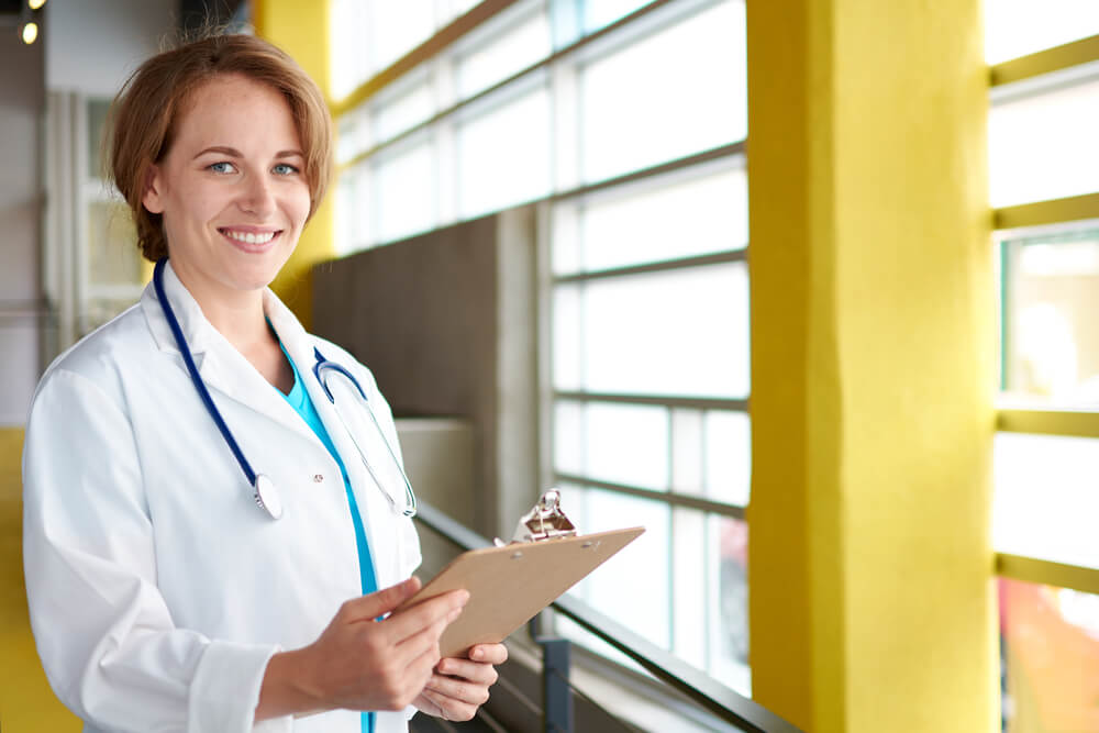 Smiling female nurse practitioner in a white lab coat taking notes by a brightly lit window