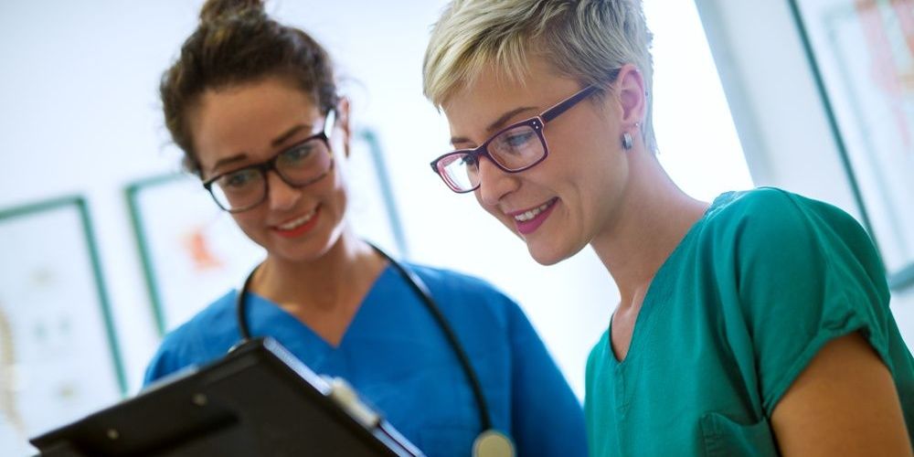 Two nurses wearing scrubs and consulting notes on a clipboard