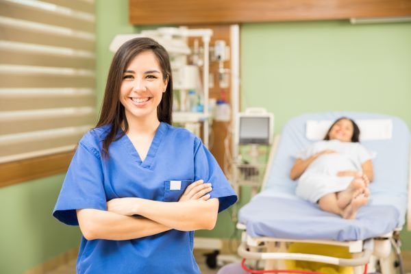 Smiling nurse in blue scrubs with a smiling patient lying on a hospital bed behind her