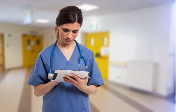 Nurse in blue scrubs using a tablet device in an empty hospital corridor