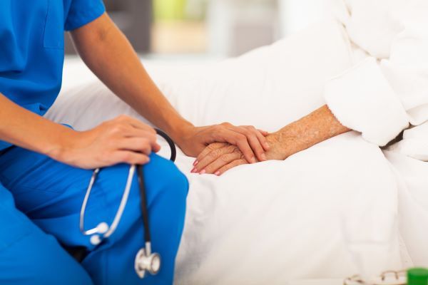 Close-up of a nurse in blue scrubs holding the hand of an elderly patient in a hospital bed