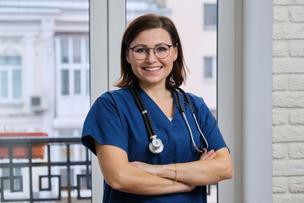 Smiling nurse in blue scrubs crossing her arms in front of a window