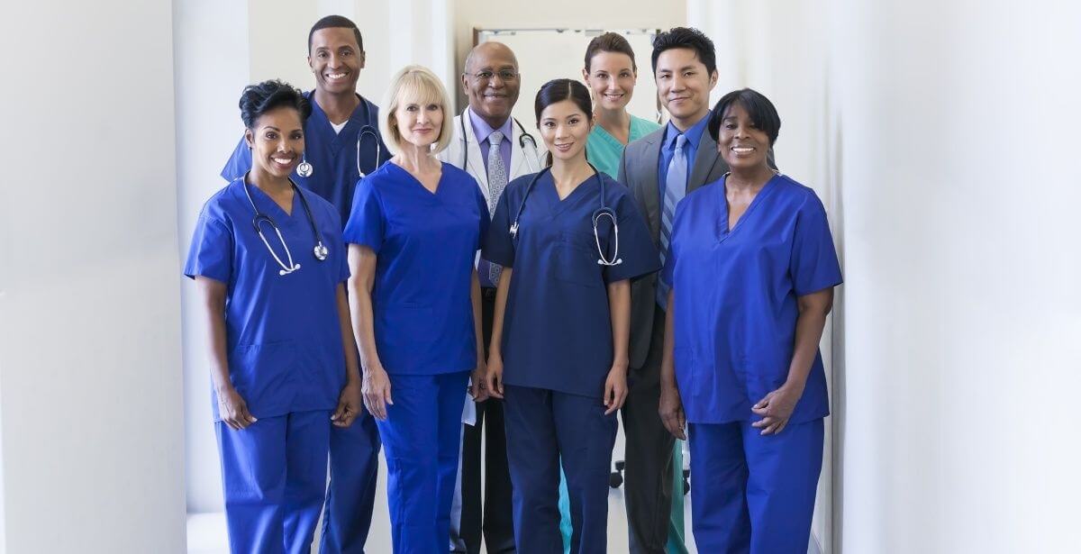 Group of ethnically diverse nurses standing and smiling in blue scrubs with other healthcare professionals